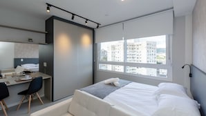 Moderni hotellihuone, jossa on sänky, työpöytä ja tuoli, suuret ikkunat, joista on näkymä kaupunkiin, sekä kattovalaisin.