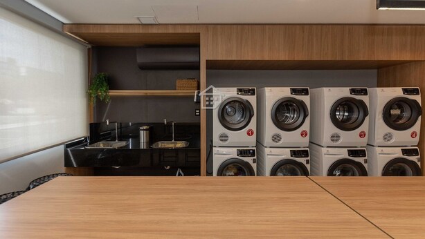 A laundry room with four stacked washing machines, a sink, and a small plant. - Studio Cubanos | Gestão Premium + Ao lado da PUC (Porto Alegre)