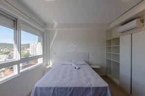 Ein Schlafzimmer mit einem Bett, einem Nachttisch, einem kleinen Tisch, einem Kleiderschrank und einem Fenster mit Blick auf die Stadt.