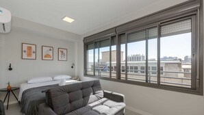 Un dormitorio moderno con un ventanal grande, un sofá gris, una cama y tres obras de arte enmarcadas en la pared.