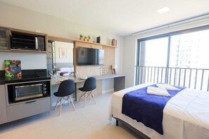 Ein kompaktes Hotelzimmer mit Küchenzeile, einem Bett mit blauer Decke, einem Schreibtisch mit Fernseher und einem Balkon mit Blick.