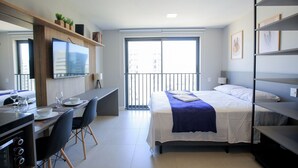 Ett modernt hotellrum med en säng, ett skrivbord, en stol, en TV och en balkong med utsikt över staden.