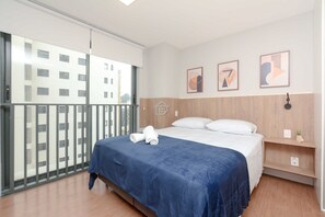 Ein Hotelzimmer mit einem Bett, einer blauen Tagesdecke, einem weißen Kissen, einem Holz-Kopfende und einem kleinen Regal mit einer Pflanze. Durch das große Fenster ist ein Gebäude zu sehen.