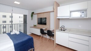 Une chambre d’hôtel moderne équipée d’un lit, d’un petit coin repas, d’une kitchenette et d’un balcon offrant une vue sur les bâtiments.
