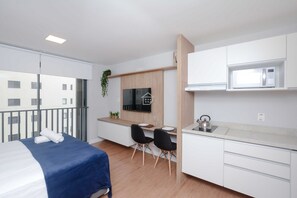 Quarto de hotel moderno com cama, pequena área de refeições, cozinha compacta e varanda com vista para edifícios.