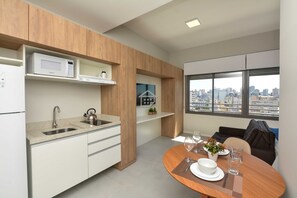 Uma cozinha moderna com micro-ondas, pia e chaleira, uma área de refeições com mesa posta para dois, e vista para a cidade atrav és de grandes janelas.