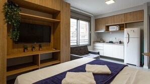 Ett modernt rum med en säng, en tv som är monterad på en träpanel, en köksdel med mikrovågsugn och kylskåp samt ett fönster.