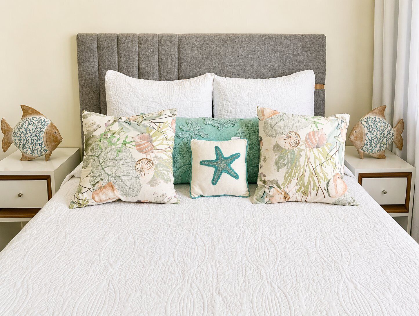 Una cama con una colcha blanca texturizada, un cabecero tapizado en gris y varios almohadones con motivos marinos.