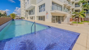 A swimming pool with blue mosaic tiles, surrounded by a multi-story building with balconies and windows. - Apartamento Delicado Próximo a la Playa (Boca Chica)
