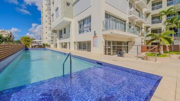 A swimming pool with blue mosaic tiles, surrounded by a multi-story building with balconies and windows.