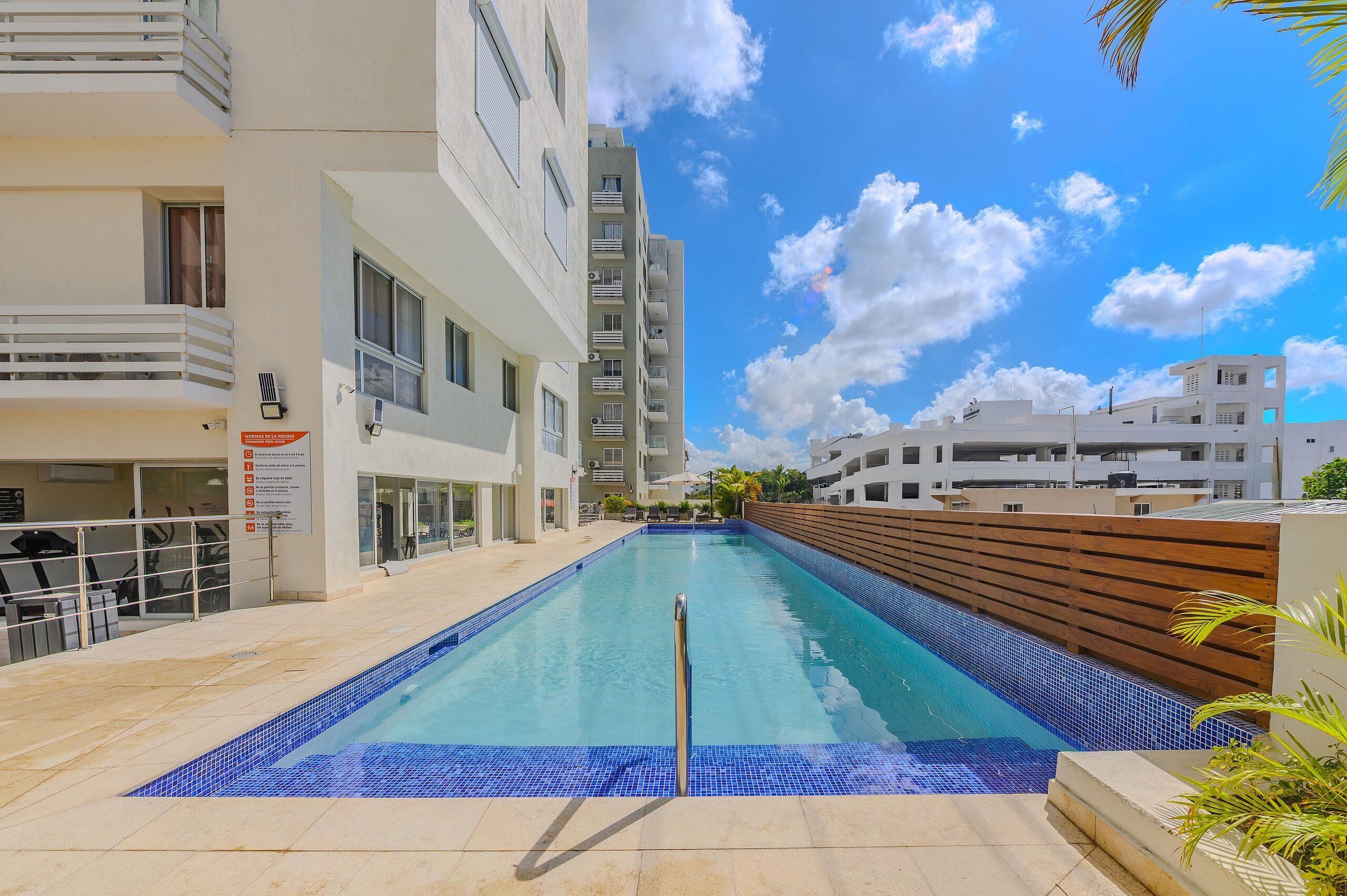 Una piscina con cubierta de madera, un edificio blanco con balcones y un cielo azul despejado.