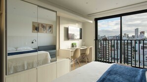 A modern hotel room with a bed, desk, and chairs, a city view, and a large window. - Studio Elegante e Conectado em Porto Alegre (Porto Alegre)