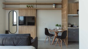 Ein modernes Hotelzimmer mit Bett, Fernseher, Essbereich mit Stühlen, einer kleinen Küche und einem großen Spiegel.