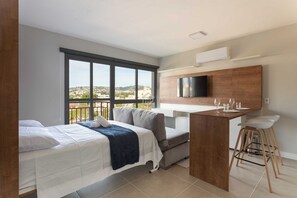 A modern hotel room with a bed, a small kitchenette, a TV, and a balcony with a city view. - Studio Minimalista e Fluido no Jardim Botânico (Porto Alegre)