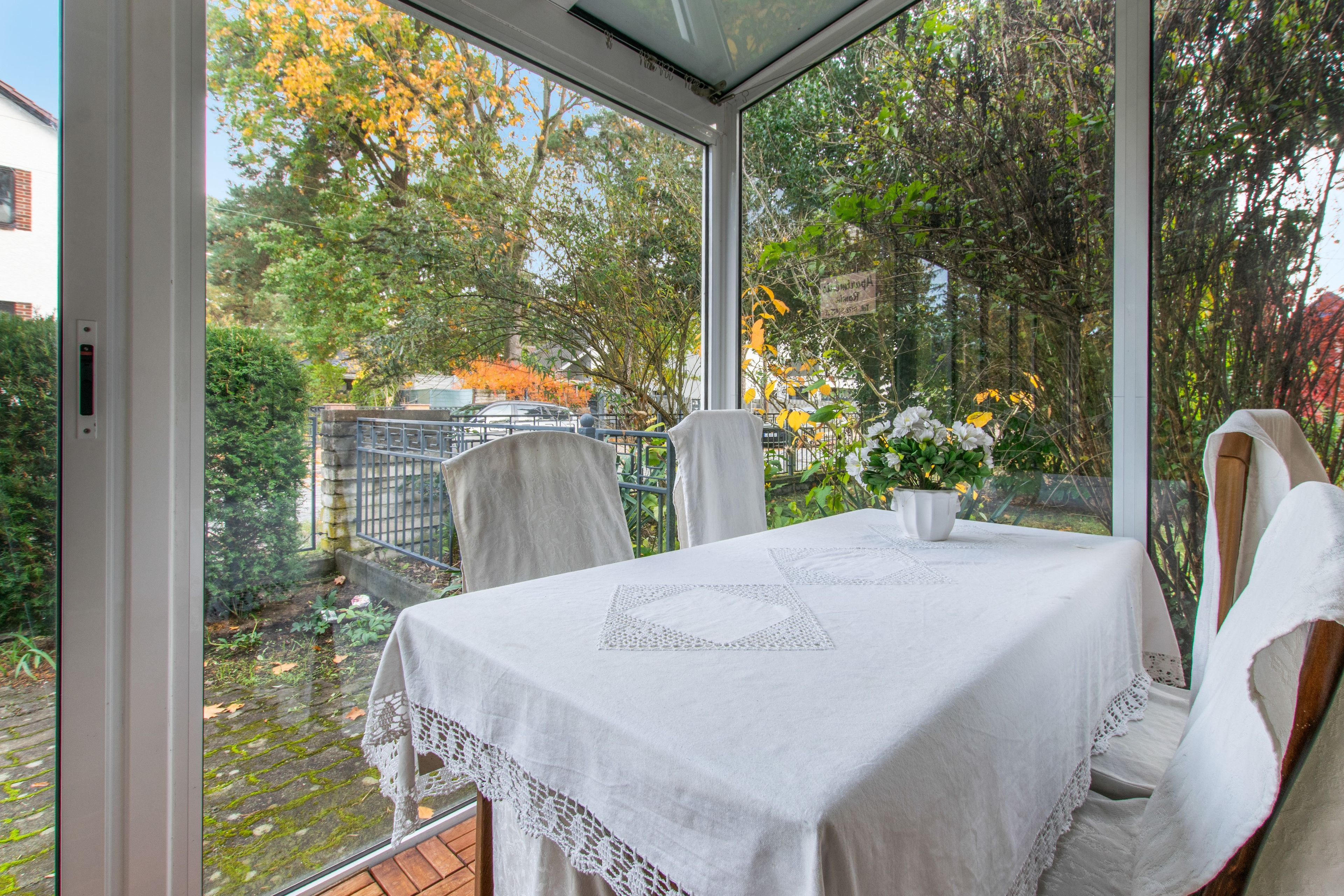Ein Wintergarten mit Tisch für zwei Personen, umgeben von Stühlen und Blick auf einen Garten mit Bäumen und einem Zaun.