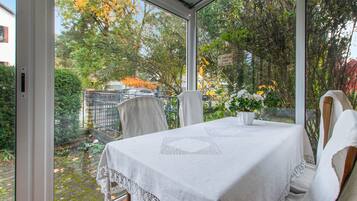 Ein Wintergarten mit Tisch für zwei Personen, umgeben von Stühlen und Blick auf einen Garten mit Bäumen und einem Zaun.
