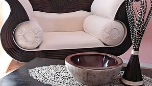 Un sofá con estructura oscura y cojines claros, un cuenco de cerámica y un florero sobre una mesa con un mantel de encaje.
