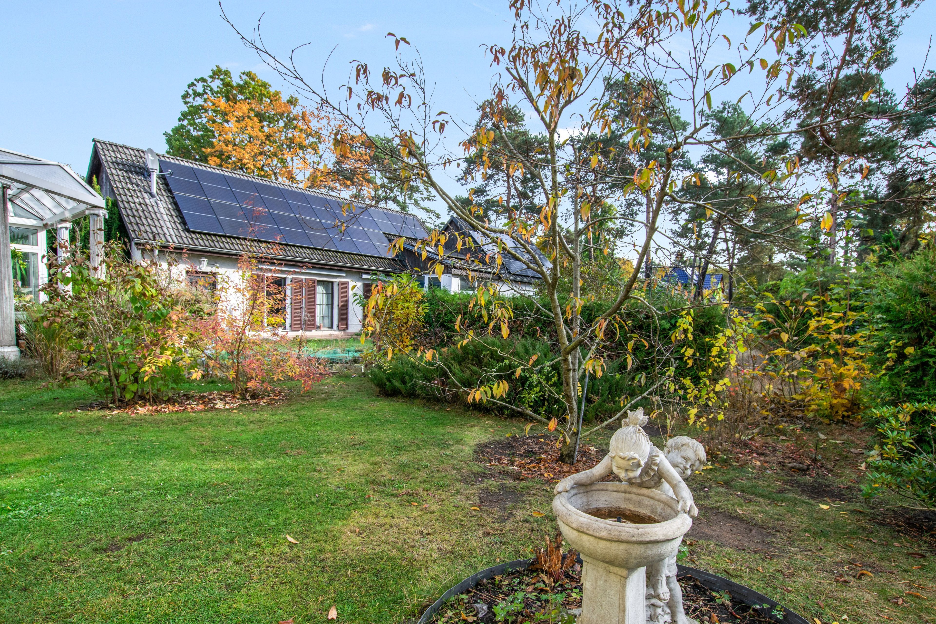 A house with solar panels, a garden with a stone fountain, and a tree with autumn leaves.