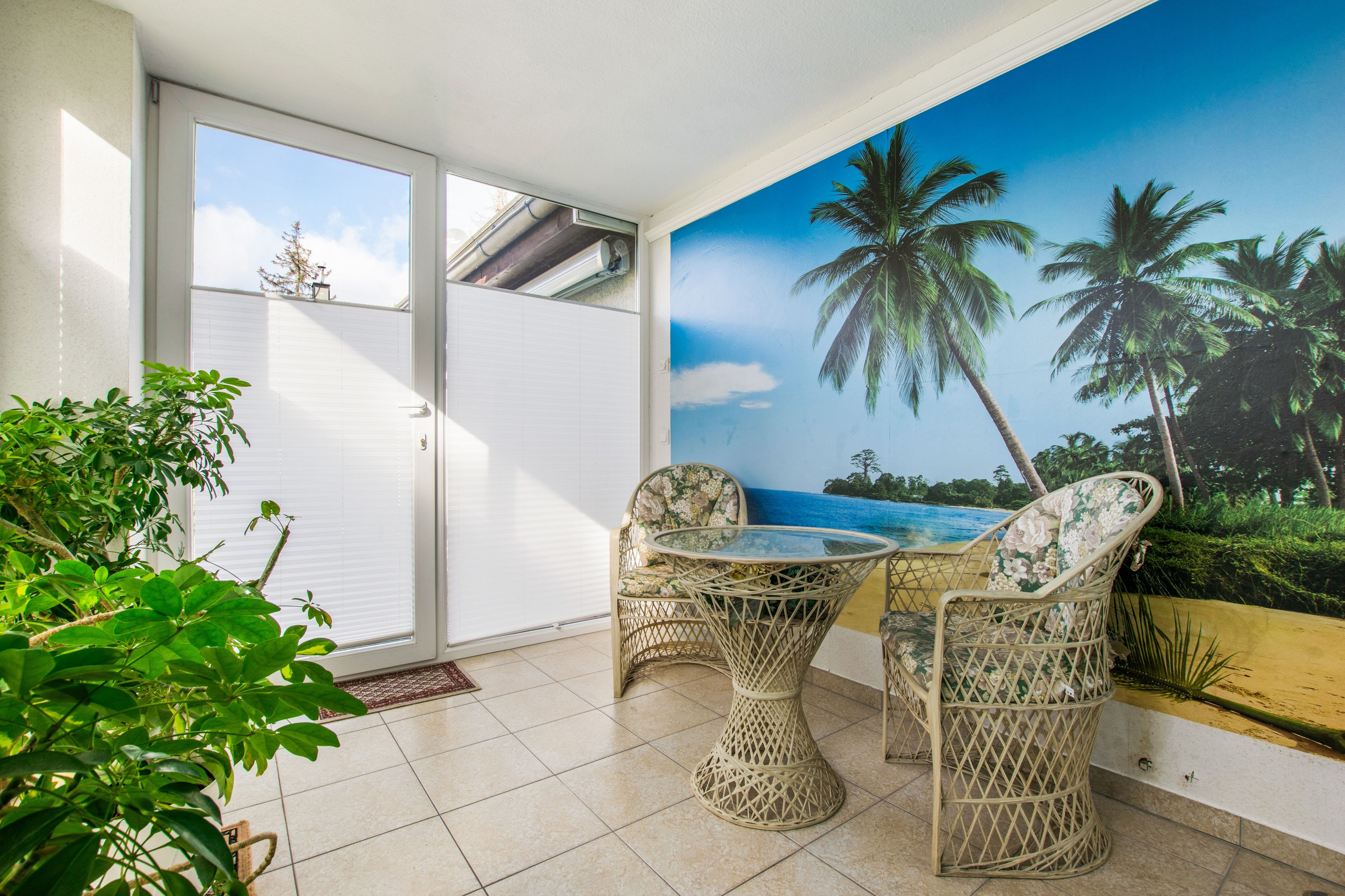 A room with a glass door leading to a balcony, a wicker table with two chairs, and a large tropical mural on the wall.