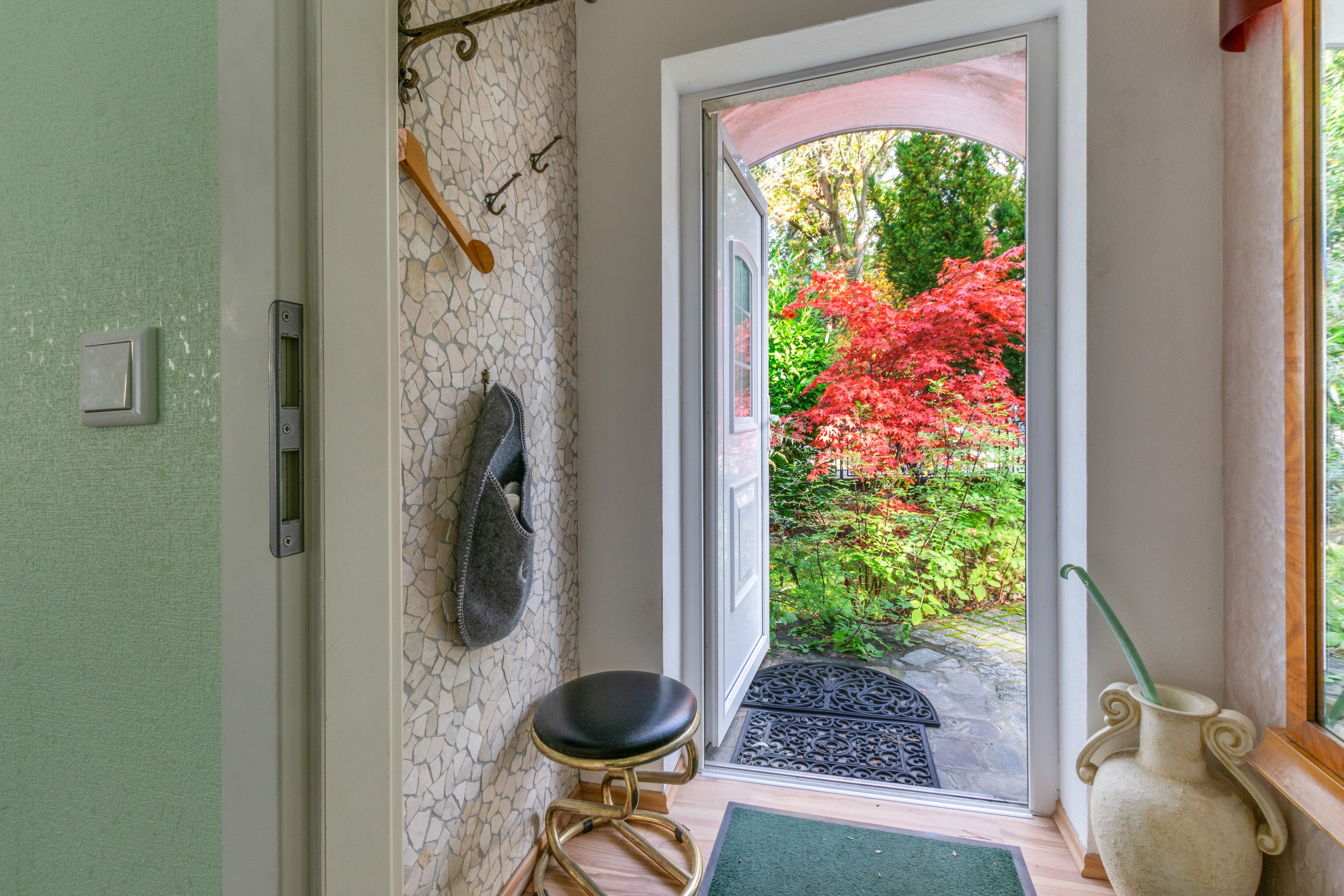 A room with a stone wall, a black leather stool, a white vase, and a view of a garden with red foliage.