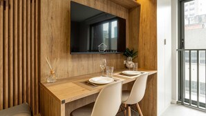 A modern dining area with a wooden table, white chairs, a flat-screen TV, and a view of the city through a large window. - Studio Moderno e Conectado em Porto Alegre (Porto Alegre)