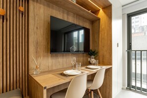Uma sala de jantar moderna com uma mesa de madeira, cadeiras brancas, uma TV de tela plana e vista para a cidade através de uma grande janela.