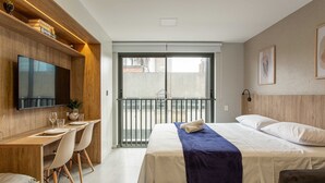 A hotel room with a bed, a desk with chairs, a TV, and a balcony with a railing. - Studio Moderno e Conectado em Porto Alegre (Porto Alegre)