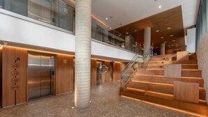 A modern hotel lobby with a central elevator, a staircase, and a glass railing on the upper level. - Studio Moderno | Lazer Completo em Porto Alegre (Porto Alegre)