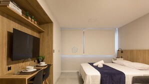 A modern hotel room with a wooden desk, a bed, a TV, and a window. - Studio moderno: rooftop no coração do Rio Branco (Porto Alegre)