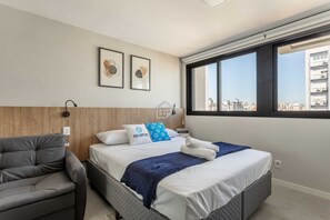 A hotel room with a bed, a grey sofa, a window with a city view, and two framed pictures on the wall. - Studio moderno no Rio Branco | Próximo à Redenção (Porto Alegre)