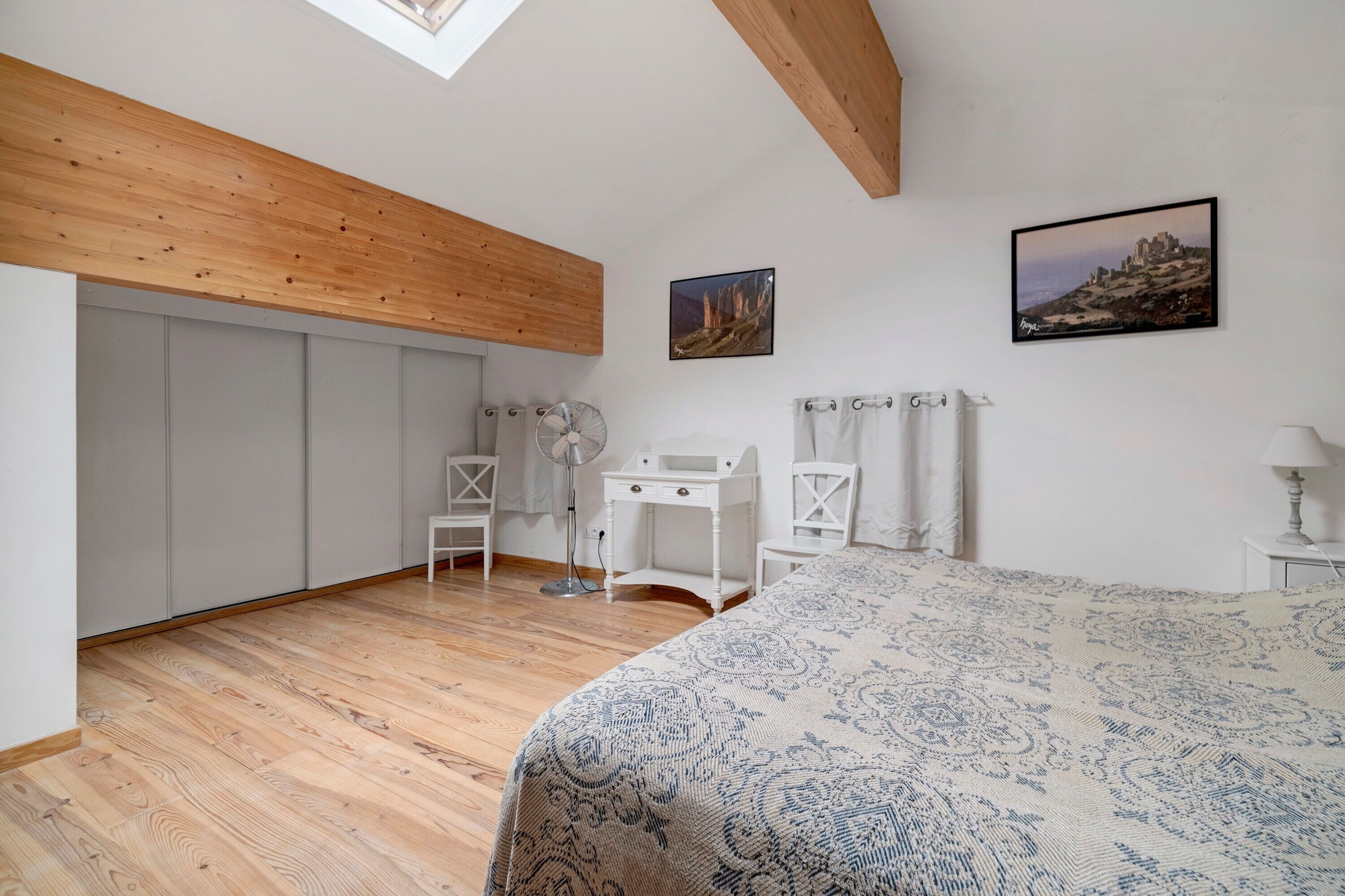 A bedroom with a bed, a desk, a chair, a fan, and a picture on the wall.