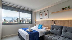 Ein modernes Schlafzimmer mit Blick auf die Stadt, ein Bett mit blauer Decke, ein graues Sofa und ein Regal mit Dekoration.