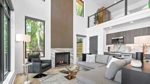 A modern living room with a fireplace, grey sofas, a glass coffee table, and a kitchen area with a microwave.