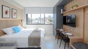 A modern hotel room with a bed, a desk with chairs, a TV, and a city view. - O Seu refúgio de estilo no coração do Moinhos (Porto Alegre)