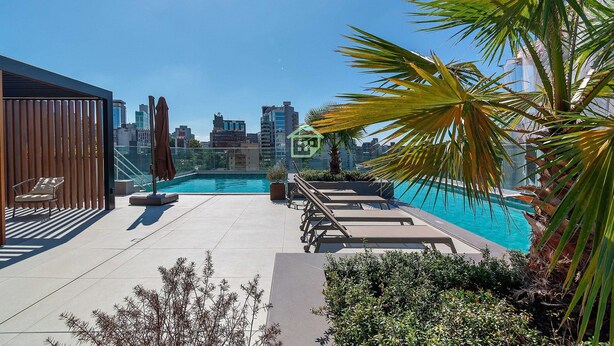 A rooftop pool area with lounge chairs, a palm tree, and a city skyline in the background. - Apto Aconchegante Próximo ao Parque Moinhos (Porto Alegre)