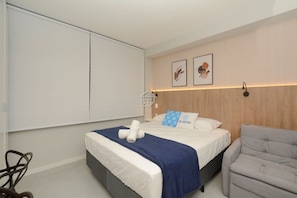 Quarto de hotel com cama, poltrona cinza, guarda-roupa e duas imagens emolduradas na parede.