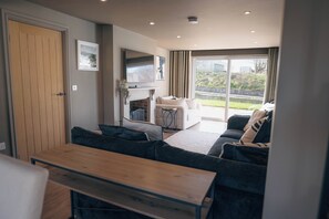 A modern living room with a fireplace, a sofa set, a dining table, and a large window offering a view of the outdoors. - Ffordd-Y-Wylan by Guestz (Rhosneigr)