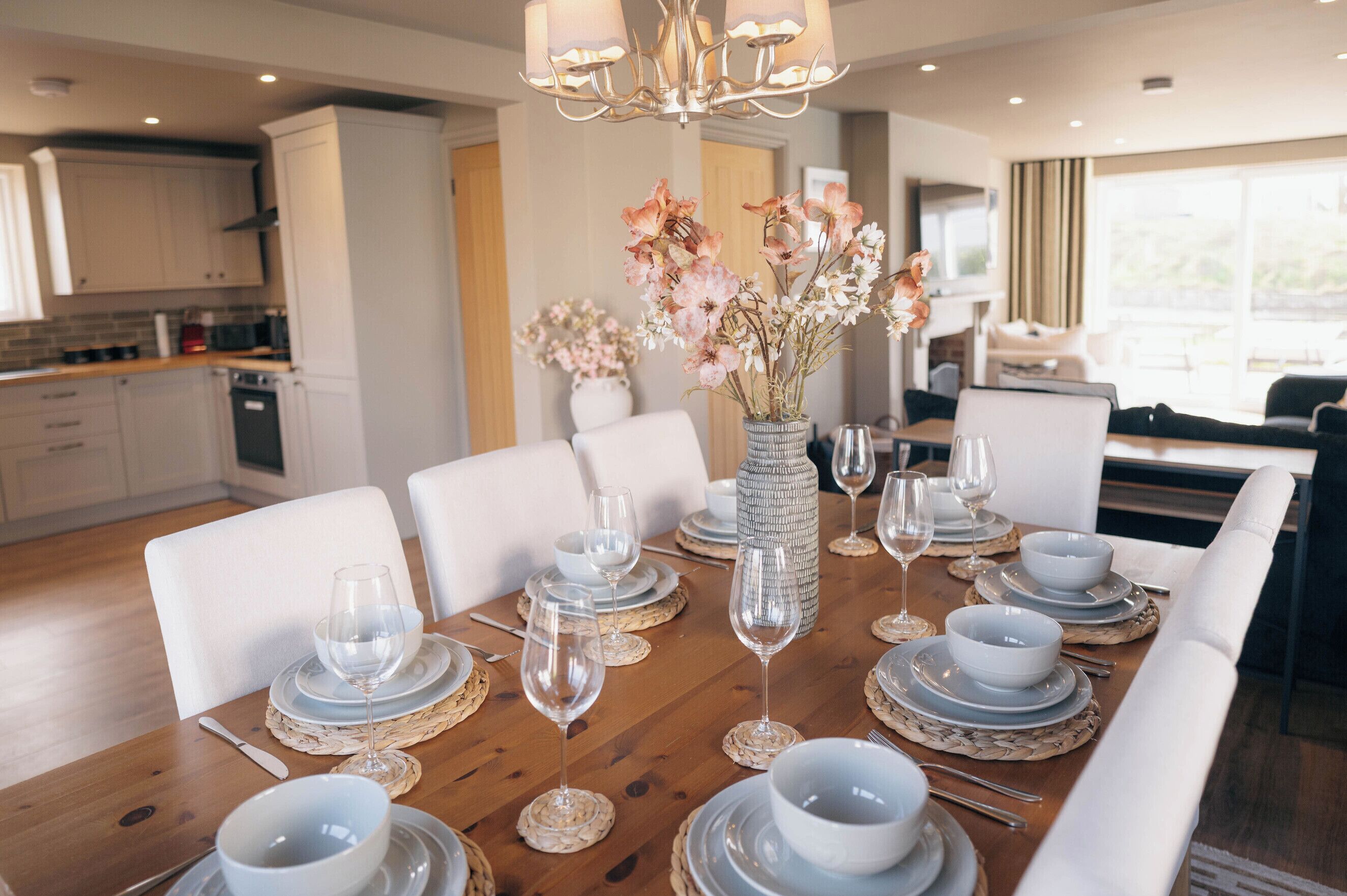 Een eetgedeelte met een houten tafel gedekt voor zes personen, witte stoelen, een vaas met bloemen en een keuken op de achtergrond.