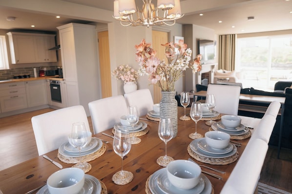 A dining area with a wooden table set for six, white chairs, a vase with flowers, and a kitchen in the background.