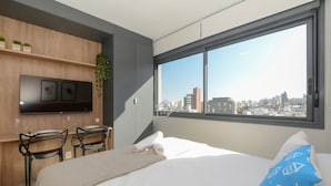 A modern hotel room with a bed, a desk with chairs, a TV, and a large window with a city view. - Sofisticação e Conforto no Coração do Moinhos (Porto Alegre)