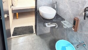 A modern bathroom with a marble wall, a white sink, a blue toilet, and a bed visible through an open door.