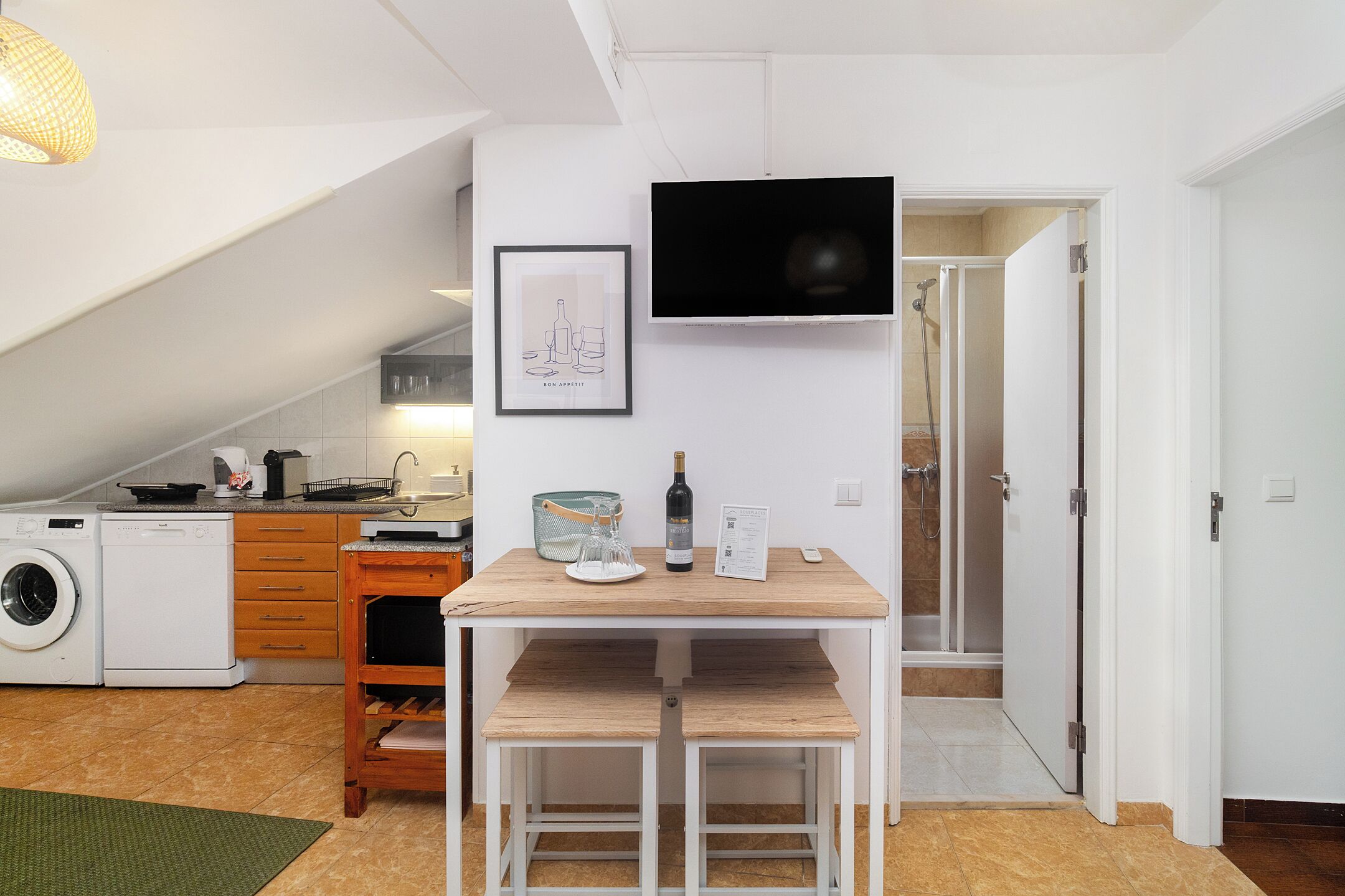 A compact kitchen with a washing machine, a dining table with chairs, a TV mounted on the wall, and a shower area.
