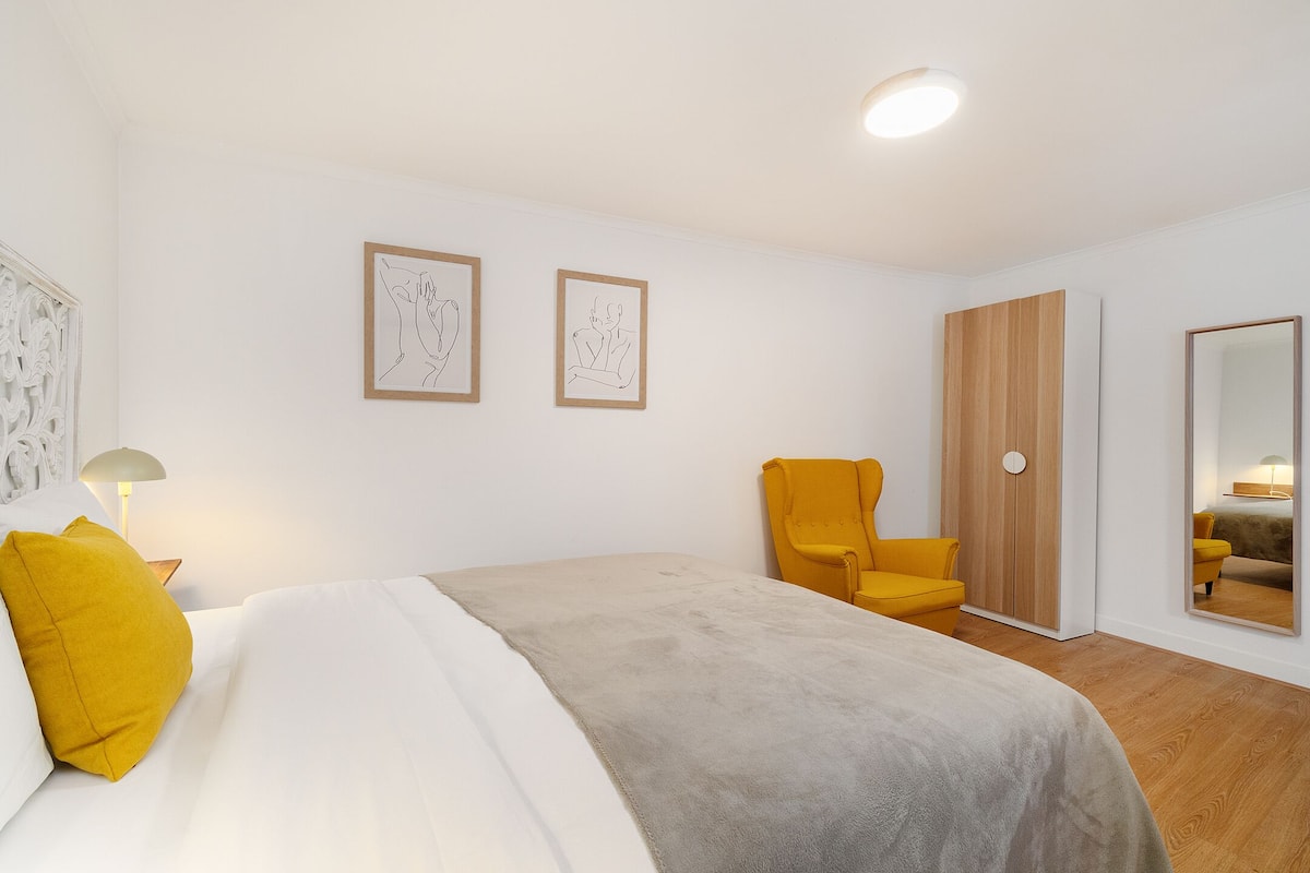 A bedroom with a bed, a chair, a wardrobe, and two framed artworks on the wall.