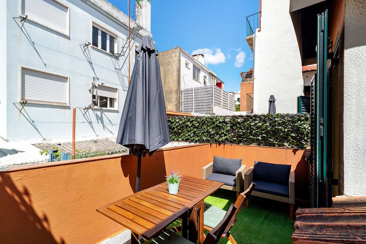 A small outdoor seating area with a table and chairs, a potted plant, and a grey umbrella.