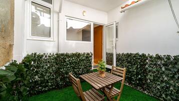 Un pequeño patio con mesa y sillas de madera, una planta en maceta y una pared blanca con ventana y puerta.