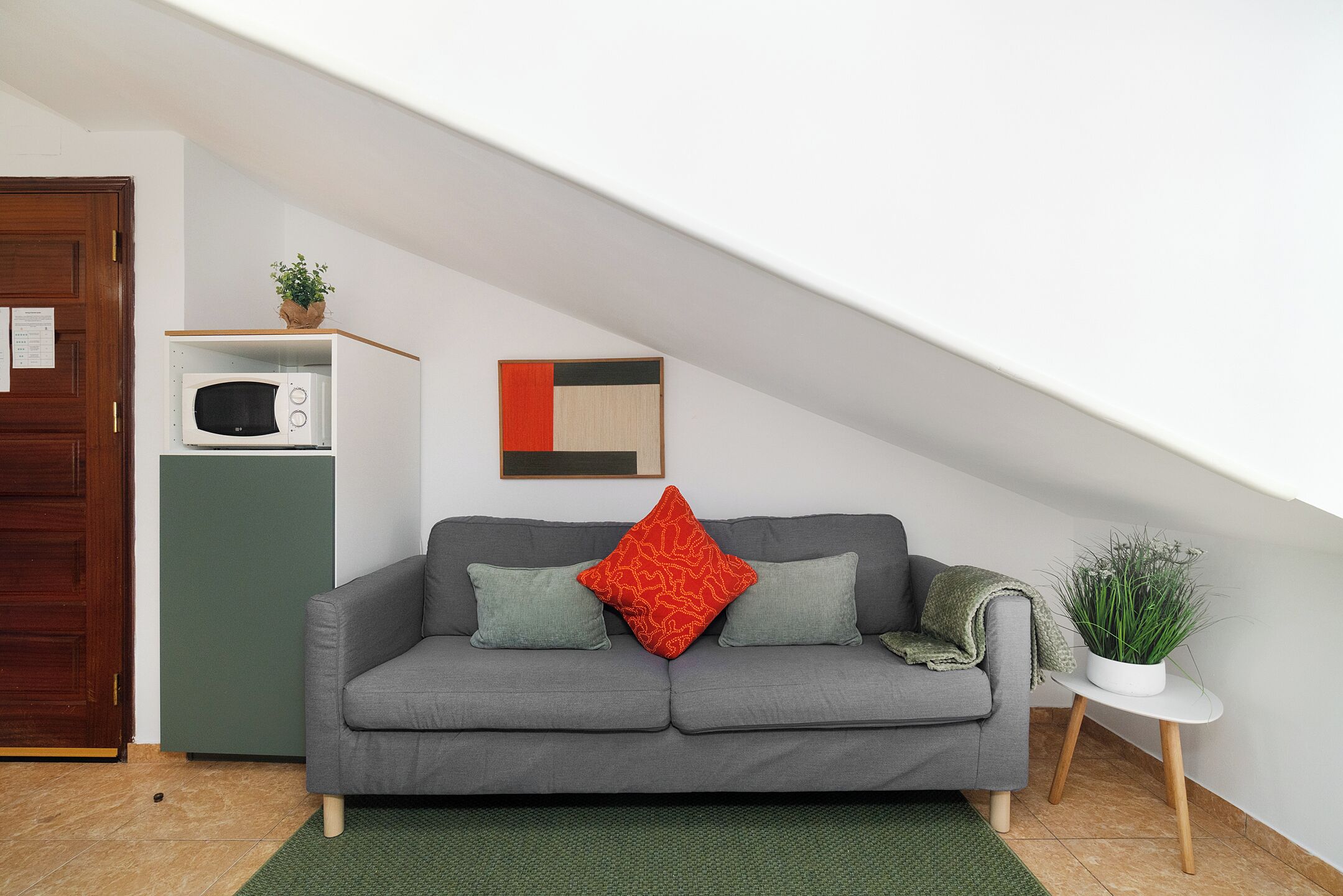 A grey sofa with two pillows, a red accent pillow, a small white table with a plant, a microwave on a shelf, and a wooden door.