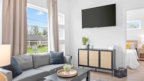 A modern living room with a grey sofa, a flat-screen TV mounted on the wall, a wooden cabinet, and a bed with white bedding.