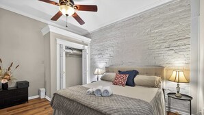 A bedroom with a bed, a ceiling fan, a nightstand with a lamp, a dresser, and a door leading to another room.
