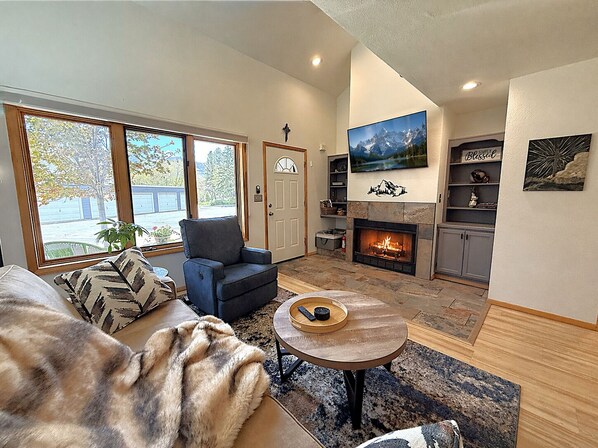 A living room with a fireplace, a television mounted above it, and a large window with a view of trees and a building.