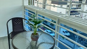 A balcony with a glass table, chairs, a potted plant, and a view of a parking lot and buildings. - Modern Apartment Comfortable & Quiet (Santo Domingo Este)